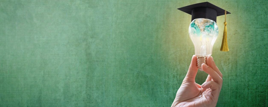 A hand holding a lightbulb with a graduation cap on top, in front of a black board